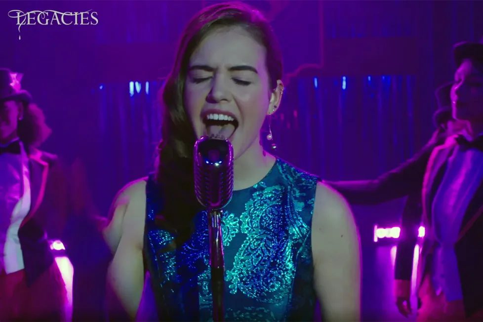 Legacies singer at a vintage microphone in purple light, with dancers in top hats behind her